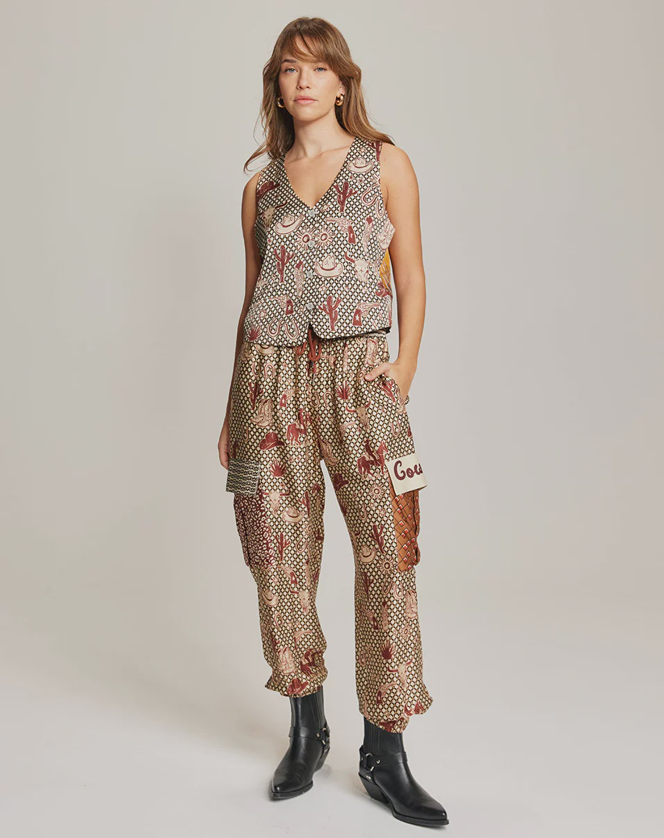 A person stands against a plain background wearing Me369's Gianna Cargo Pants, featuring a high-waisted Rodeo print and "Gou" patch, paired with a sleeveless patterned top and black ankle boots, with one hand in their pocket.