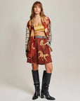 A woman wears the Me369 Juliette Pleated Shorts—a red and gold patterned set with an elastic waist—paired with a yellow crop top, checked belt, black knee-high boots, and loose brown hair as she faces the camera neutrally.