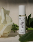 A white MAYA NO.2 10ML bottle by Maya Fragrances, featuring a rose illustration, stands upright beside a white rose and green leaves on a neutral background, evoking the sexy, musky undertones of this perfume oil.
