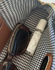 A close-up of a brown bag with black and white houndstooth lining holds tortoiseshell sunglasses and a bottle of Maya Fragrances' MAYA BASE 10 vegan, cruelty-free fragrance oil.