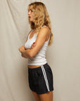A person with long wavy blonde hair, in a white tank top and Perfectwhitetee Mikayla Travel Short—ultra-comfy dark athletic shorts with white stripes—stands in profile, arms crossed against a plain light backdrop.