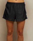 A person faces forward, wearing Perfectwhitetee's Mikayla Travel Short in dark gray with white side stripes. These ultra-comfy shorts feature an elastic waistband, and the person's midriff and legs are visible against a neutral background.