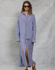 A woman in sunglasses and green sandals wears the Mamene Slim Phoebe Dress, a blue-and-white striped cotton shirtdress, standing against a light gray textured backdrop with one leg slightly forward.