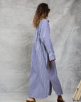 A woman with curly hair and sunglasses walks on a wooden floor against a light backdrop, wearing Mamene’s Slim Phoebe Dress—an ankle-length, navy and white striped cotton shirtdress—with green wedge sandals.