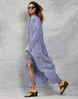 A woman with curly hair and sunglasses wears the Mamene Slim Phoebe Dress—an ankle-length navy and white striped cotton shirtdress—and green wedge sandals as she walks confidently before a gray textured backdrop.