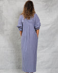 A person with curly hair faces away, wearing Mamene's Slim Phoebe Dress—an ankle-length, navy and white striped cotton shirtdress with rolled-up sleeves—and olive green heeled sandals against a textured light gray background.