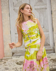A woman poses by a rustic wooden door, wearing the Hunter Bell NYC Fallon Skirt, featuring colorful green, pink, and yellow abstract patterns. The midi-length linen-rayon blend flows gracefully with pleats as her long hair cascades down her back. She looks to the side with a relaxed expression.