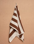 The Maria Hand Towel by Autumn Sonata, featuring brown and white stripes, hangs on a hook against a tan wall. Crafted from soft organic cotton, it offers everyday comfort and style.