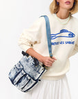 A woman wears a cream sweatshirt with a blue fish graphic and "LIBEREZ LES SARDINES" text, a white pleated skirt, and carries a blue denim handbag featuring the Clare Vivier Adjustable Crossbody Strap over her shoulder.