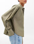 A person wears the Rag & Bone martha poplin striped shirt, a boxy fit olive-green top with white vertical stripes, paired with light blue high-waisted jeans against a plain white background.