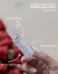 A hand holds a MAYA BASE 10 roll-on by Maya Fragrances. Arrows highlight lily petals, sandalwood, amber, and soft musk notes. Red flowers blur behind. Text reads “THE NOTES maya base.” Vegan and cruelty-free.