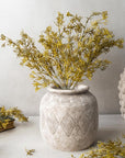 The Abigail Ahern Meadow Grass Flaxen is arranged in a textured ceramic vase with a diamond pattern, displayed on a light surface beside similar decor and foliage against a neutral background.