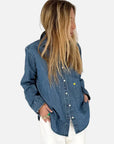 A person with long blonde hair wears the Kerri Rosenthal Mia Denim Shirt—an oversized denim top with an embroidered heart—paired with white pants, standing against a plain light background and looking to the side, one hand in their pocket.