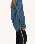 A person wearing the Kerri Rosenthal Mia Denim Shirt, featuring an embroidered heart patch on the elbow, paired with black pants with a white side stripe, stands sideways against a plain background.