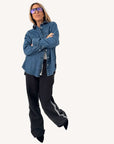 Wearing a Kerri Rosenthal Mia Denim Shirt with an embroidered heart, blue-tinted glasses, black wide-leg pants with side detailing, and black pointed-toe shoes, a person stands arms crossed against a plain background.