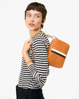 A woman in a black-and-white striped long-sleeve shirt and black pants holds the Clare Vivier Midi Sac S25, a handwoven leather rectangular crossbody with a gold chain strap, resting it on her shoulder and looking to the side.