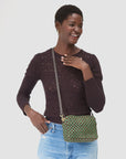 A smiling woman in a dark, textured long-sleeve top and light blue jeans holds the Clare Vivier Midi Sac 26, a handwoven green and brown checkered leather crossbody bag, standing against a plain white background.