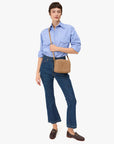 A person with short brown hair and a neutral expression poses against a white background, wearing a light blue button-up shirt, blue jeans, brown loafers, and carrying the Clare Vivier Midi Sac S25 tan rectangular crossbody bag.