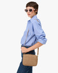 Wearing a light blue button-up shirt, dark jeans, and pink cat-eye sunglasses, a person stands sideways holding the Clare Vivier Midi Sac S25—a rectangular crossbody bag with a long detachable strap and handwoven leather.