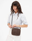 A woman with long wavy brown hair wears a light blue button-up shirt and white pants, holding the Clare Vivier Midi Sac S25 crossbody bag with a black strap against a plain white background.
