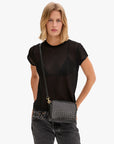 A person with shoulder-length blonde hair wears a sheer black t-shirt, dark jeans, and carries the Clare Vivier Midi Sac S25 crossbody bag with a woven pattern, standing against a plain white background.