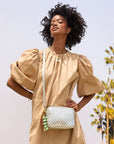 A woman in a beige puff-sleeve dress poses outdoors with the Clare Vivier Midi Sac S25, a chic handwoven leather crossbody bag. Palm trees and a clear sky are behind her. She has curly hair and wears layered necklaces.