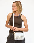 A woman with wavy blonde hair, in a sleeveless dark brown top and black jeans, holds the Clare Vivier Midi Sac S25 handwoven leather crossbody bag. She wears a purple beaded necklace and looks off to the side against a plain background.