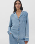 A woman with long dark hair wears the Rag & Bone miramar slink long sleeve lounge shirt and matching denim pants, standing against a plain white background, one hand in her pocket.