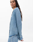 A woman with long dark hair wears a Rag & Bone miramar slink long sleeve lounge shirt and matching denim-print pants, standing against a plain white background and glancing over her shoulder.