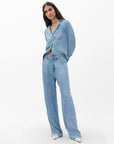 A woman with long dark hair wears Rag & Bone’s miramar slink lounge pant in a trompe l’oeil denim print with a matching shirt, both crafted from soft loungewear fabric, against a plain white background. She finishes the look with pointed silver shoes.