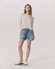 A person with long hair stands against a plain background in a light, long-sleeve shirt and white loafers, wearing Rag & Bone's Miramar Walking Short featuring denim print and an elastic waistband. They strike a relaxed pose.