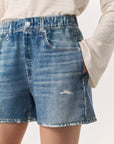 A person is wearing Rag & Bone's Miramar Walking Short with frayed and distressed denim print, paired with a cozy light-colored long-sleeve top. With one hand casually in their pocket, the look seamlessly blends casual flair with style.