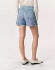 The person stands on a light floor wearing a long-sleeved shirt, Rag & Bone's Miramar Walking Short featuring denim print, frayed hems, and rear pockets, paired with white loafers against a plain backdrop.