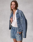 A person with long hair stands against a neutral gray background, wearing Rag & Bone's Miramar Walking Short styled as denim print shorts, paired with a denim jacket and white shirt, looking directly at the camera with a relaxed expression.
