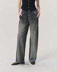 A person stands against a plain light background with hands in pockets, wearing Rag & Bone’s Miramar Wide Leg Track Pant in faded black denim look, a black sleeveless buttoned top, and black pointed-toe flats.