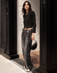 A woman with wavy dark hair stands in a doorway against white brick and ornate black columns, wearing Rag & Bone's Miramar Wide Leg Track Pant, a cropped black jacket, black sandals, and holding a small black handbag.