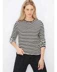 A person with long blonde hair wears "The Modern Long" black and white striped shirt by Kule, crafted from luxurious Portuguese cotton, paired with black pants. They stand against a white backdrop, holding the shirt's hem with one hand.