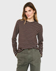 A person with light brown hair models Kule's The Modern Long, a brown and white striped long sleeve tee, paired with olive green pants, standing hands-in-pockets against a plain light gray background.