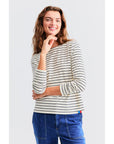 A smiling person with wavy hair models "The Modern Long" by Kule, a black and white striped long-sleeve shirt crafted from Portuguese cotton, paired with blue jeans, against a light gray background. One hand rests on their chin.