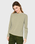 A woman with long brown hair wears Kule's The Modern Long, a green and white striped long sleeve tee, paired with green pants. She looks down and slightly smiles against a plain light gray background.