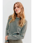 A person with long, wavy hair is wearing The Modern Long by Kule, a Portuguese cotton striped long-sleeve shirt, paired with green pants against a plain white background.