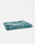 The Morelia Beach Towel by Coyuchi Inc, crafted in soft velour terry, features a teal organic fabric with a geometric pattern of white and light brown lines and rectangles. It is shown folded on a plain white background.