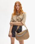 A person with long red hair wears a beige button-up shirt and a black leather skirt, holding the Clare Vivier Moyen Messenger S25, a handwoven leather handbag with a crossbody strap. The background is plain white.