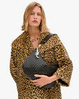 A woman in a leopard print coat holds the Clare Vivier Moyen Messenger S25, a large handwoven leather crossbody bag. She has shoulder-length blonde hair, wears a multi-pendant necklace, and stands against a plain white background.