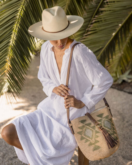 A woman in a white dress and Theo hat by Ninakuru sits under a palm tree near a Scottsdale, Arizona bungalow, holding a straw bag with green leaf patterns.