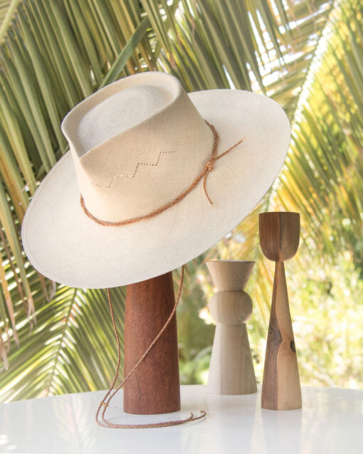 A traditional white Ninakuru Luna hat rests on a tall, wooden stand, accompanied by two minimalist wooden sculptures, set against a backdrop of lush palm leaves.
