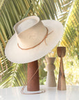 A traditional white Ninakuru Luna hat rests on a tall, wooden stand, accompanied by two minimalist wooden sculptures, set against a backdrop of lush palm leaves.