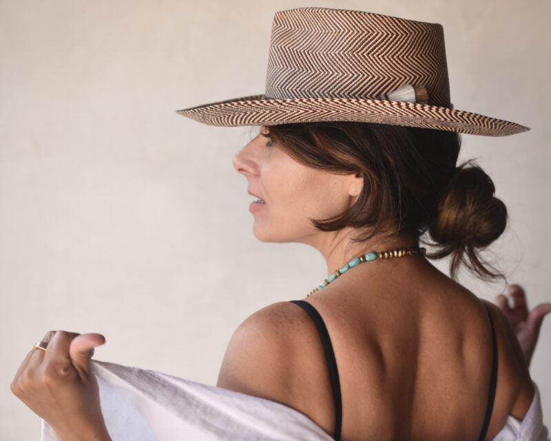 A woman in profile, wearing a stylish chevron-patterned Ninakuru Emery XLong Brim straw hat and a black tank top, glances over her shoulder while holding a white shawl. A turquoise necklace complements her ensemble.