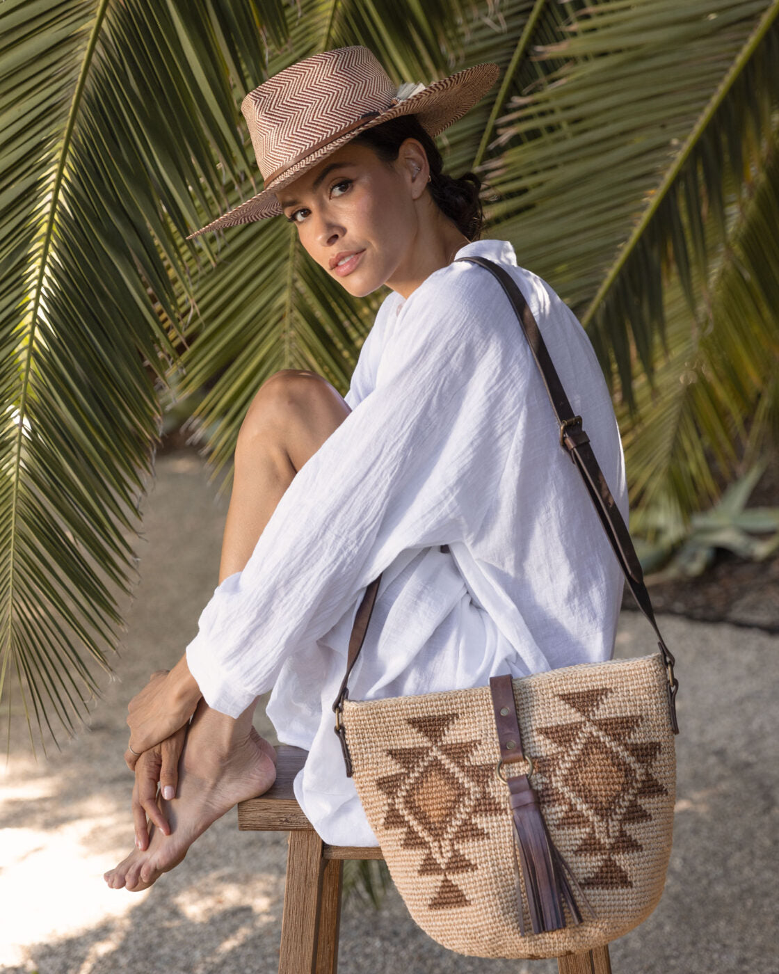 A woman sits on a wooden stool outdoors, wearing a white blouse, a straw hat (Emery XLong Brim by Ninakuru), and carrying a large woven bag with geometric designs. Tropical palm leaves form the backdrop.