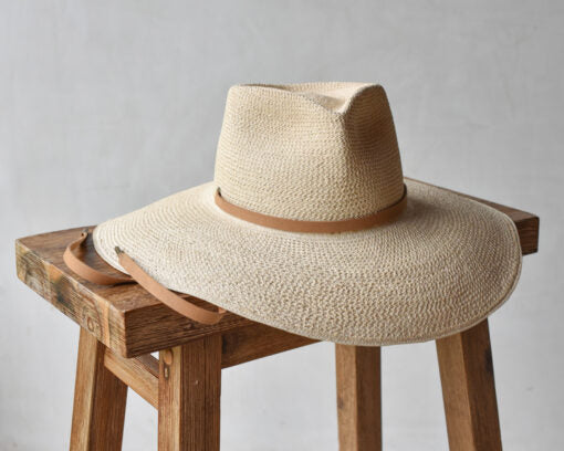 A Ninakuru Ember hat with a broad brim and a leather band rests on a wooden stool against a plain, light gray background.
